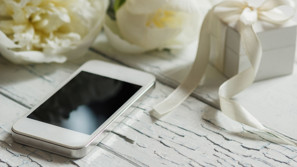 An iphone on a white-washed table with white flowers and a white candle.